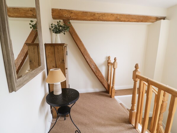 Une pièce avec un escalier en bois, une table ronde noire surmontée d’une lampe, et un miroir accroché au mur.