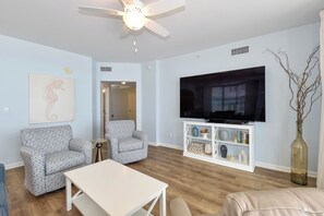 A living room with a ceiling fan, a flat-screen TV, two patterned armchairs, a white coffee table, and a bookshelf with decorative items. - Corner 6th Floor 3BR Condo at Silver Beach Towers! (Destin)