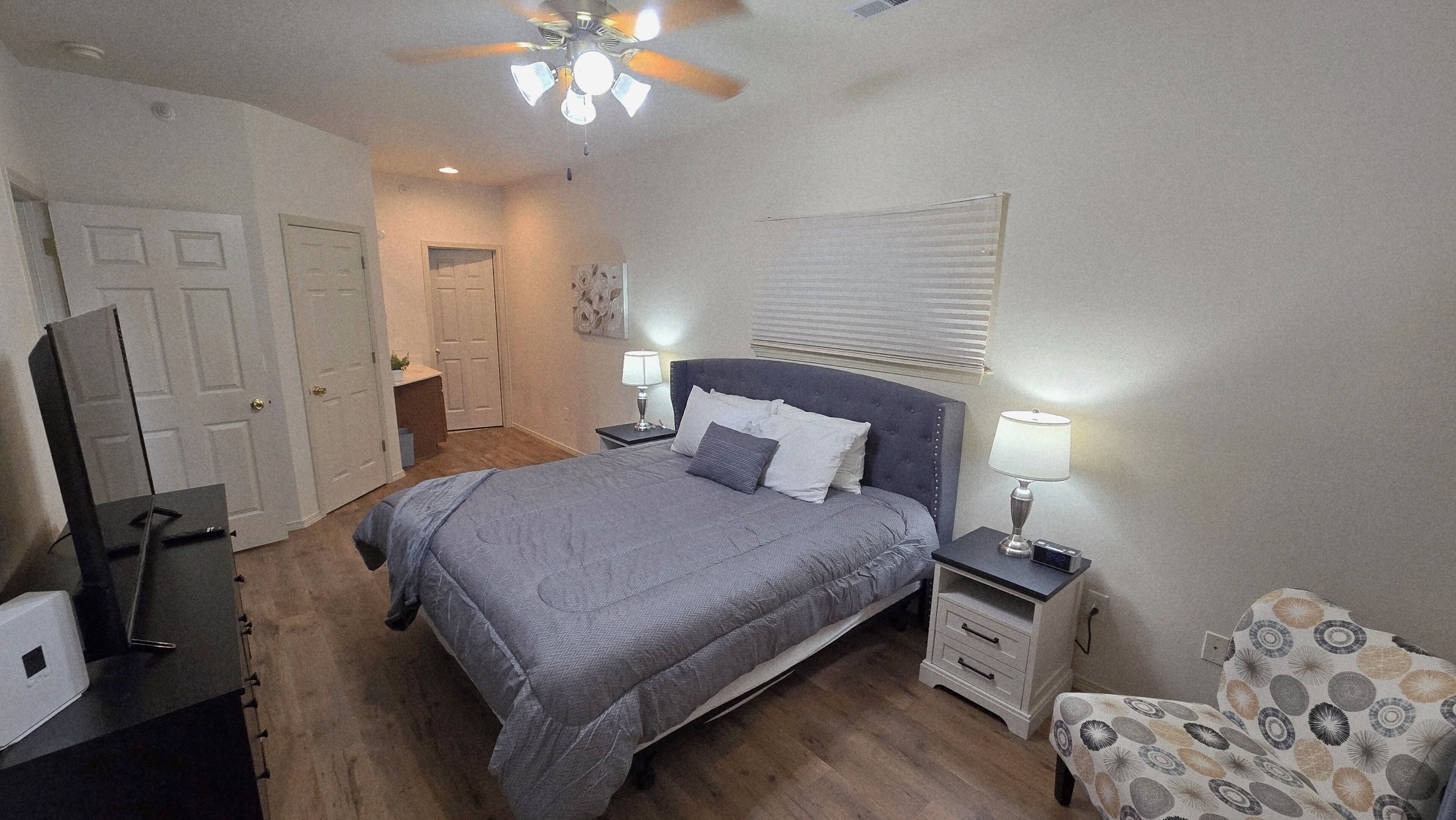 Une chambre à coucher avec un lit, des tables de chevet, une chaise, un ventilateur de plafond et une porte.