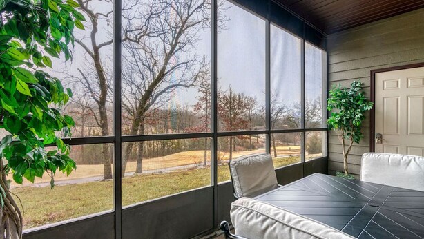 Un patio cubierto con mesa y sillas, ventanas amplias y vistas a un área verde con árboles.