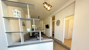 En modern inredning med en central, vit hyllsektion, en matplats med bord och stolar, ett kök i bakgrunden och en klocka på väggen.