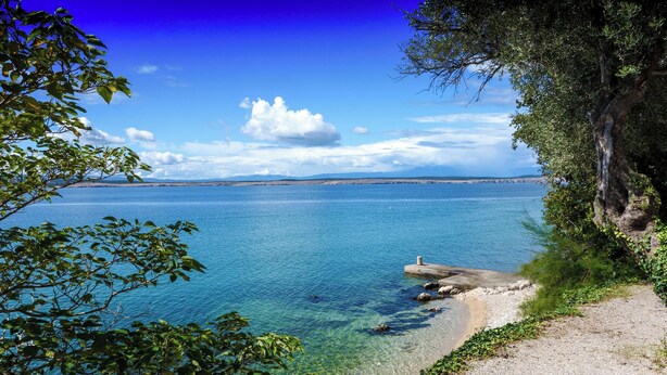 Ett kustlandskap med klart vatten, en stenig strand och en stig som leder ner till stranden.