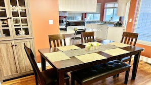 Comedor con mesa de madera preparada para cuatro personas, rodeada de sillas. Se observa una cocina al fondo.