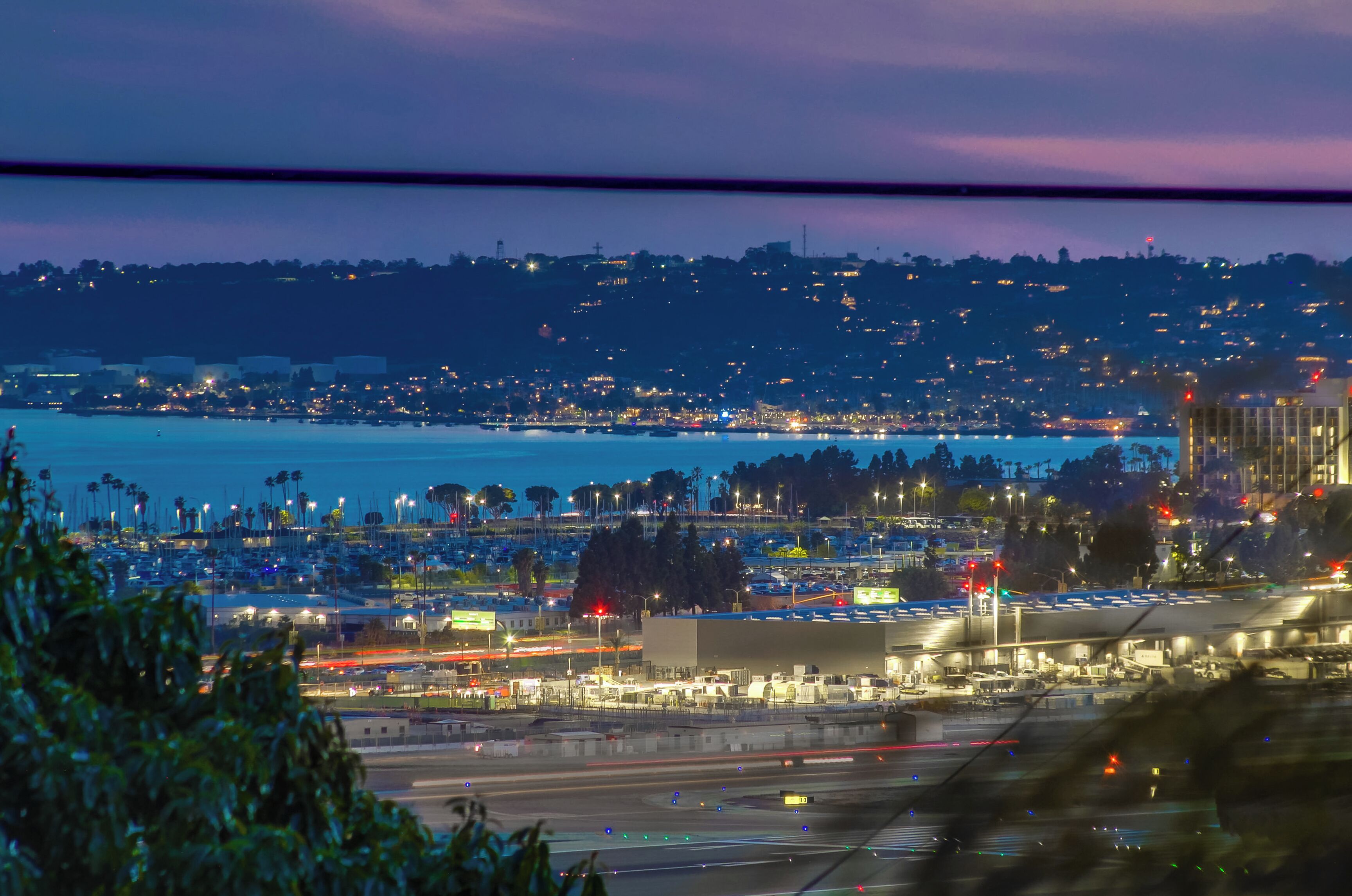 A view of a marina with boats docked, a large building with a flat roof, and a distant cityscape with lights.