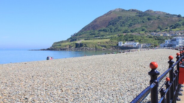 A pebbled beach with a rocky shoreline, a metal railing with red balls, and a hill with buildings in the background. - Churchmount Holiday Home Roundwood (Roundwood)