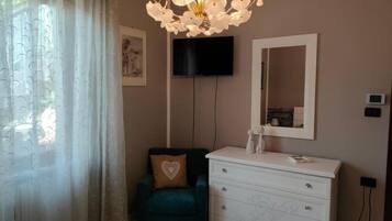 Une chambre à coucher avec un lit, un fauteuil vert, une commode blanche, un téléviseur fixé au mur et un lustre.