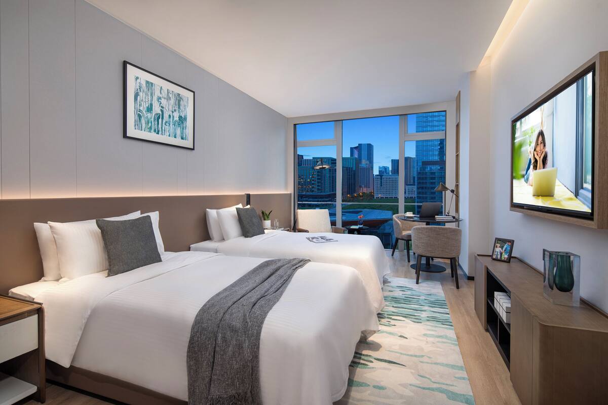 A hotel room with two beds, a desk, a chair, a TV, and a large window with a city view.