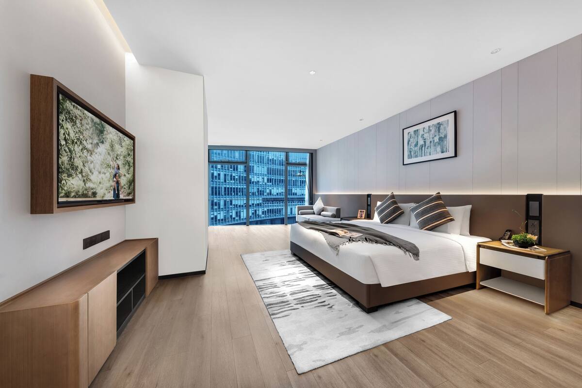 A modern bedroom with a large bed, a bedside table, a built-in TV, and a large window with a city view.