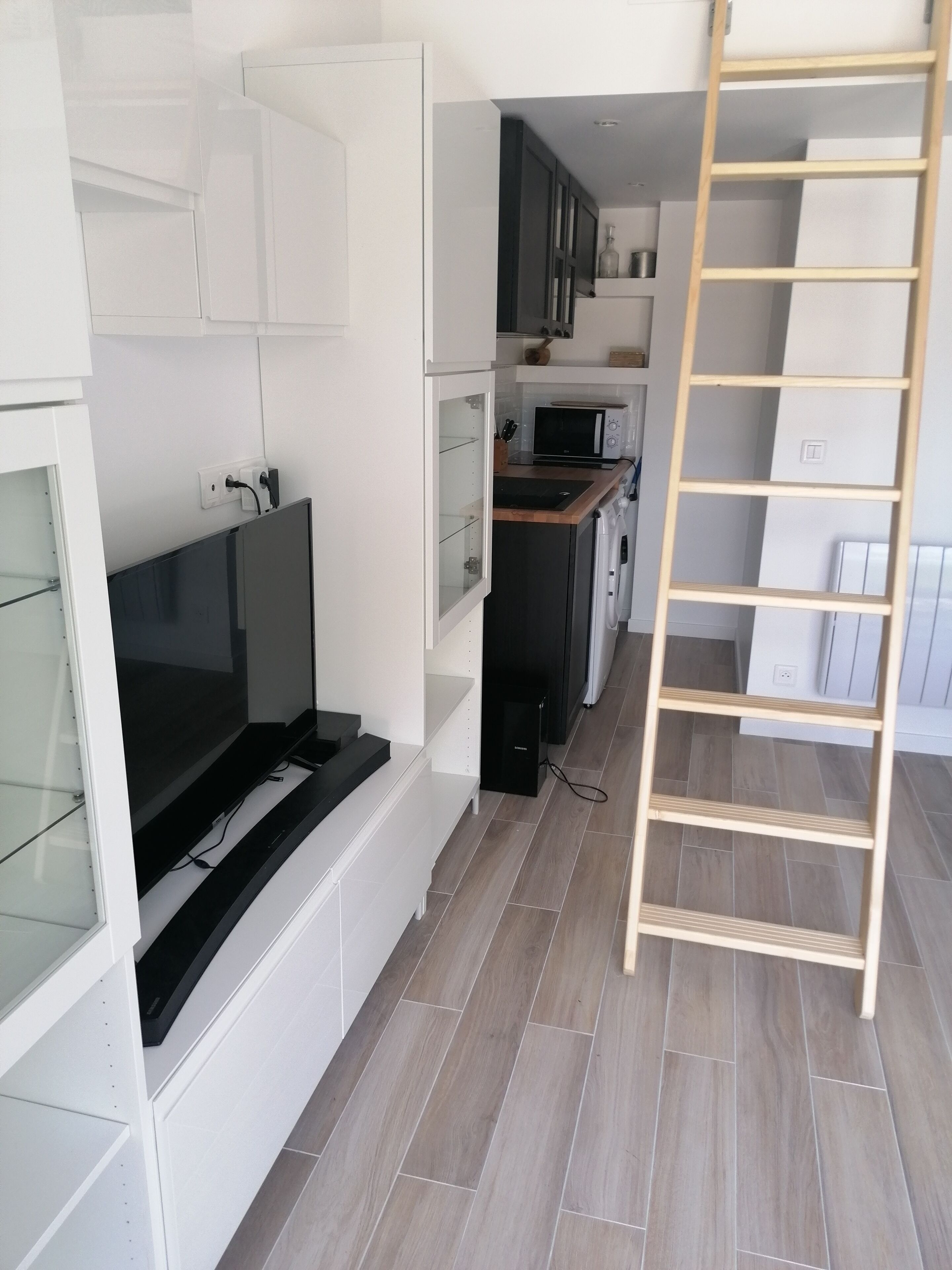 Une cuisine moderne avec des meubles blancs, un four encastré et un four à micro-ondes. Une échelle en bois est adossée au mur, et un téléviseur est fixé au mur.