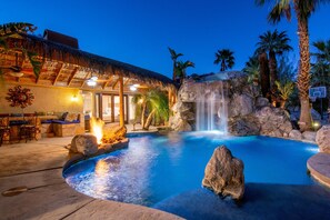 A pool with a waterfall, surrounded by rocks and palm trees, with a thatched-roof building nearby. - The Grotto- 5bd/4ba-Waterfalls-Pool-Spa-Games Room-Secluded Acreage (Desert Hot Springs)