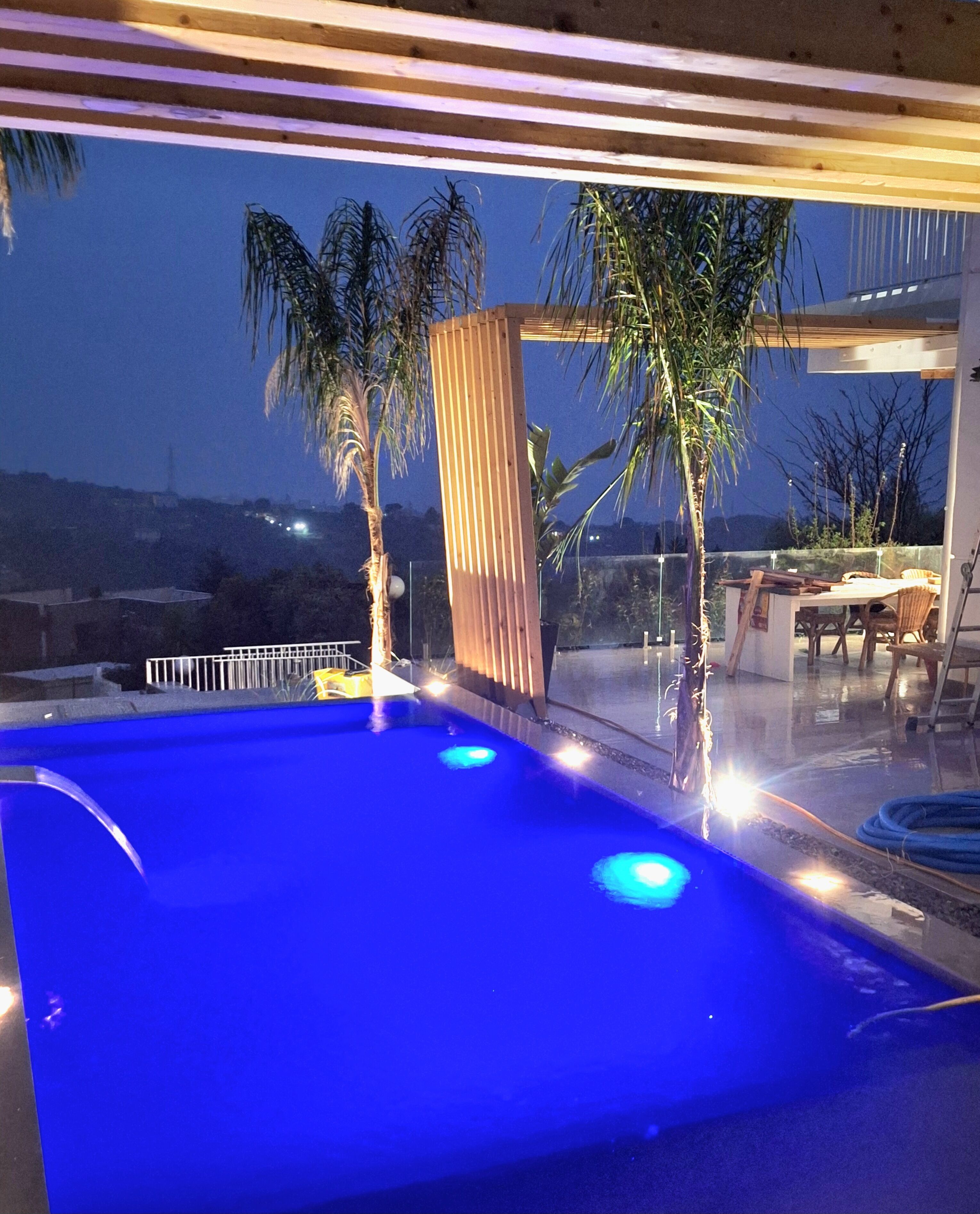 An outdoor swimming pool with blue lighting, surrounded by palm trees and a view of the cityscape at dusk.