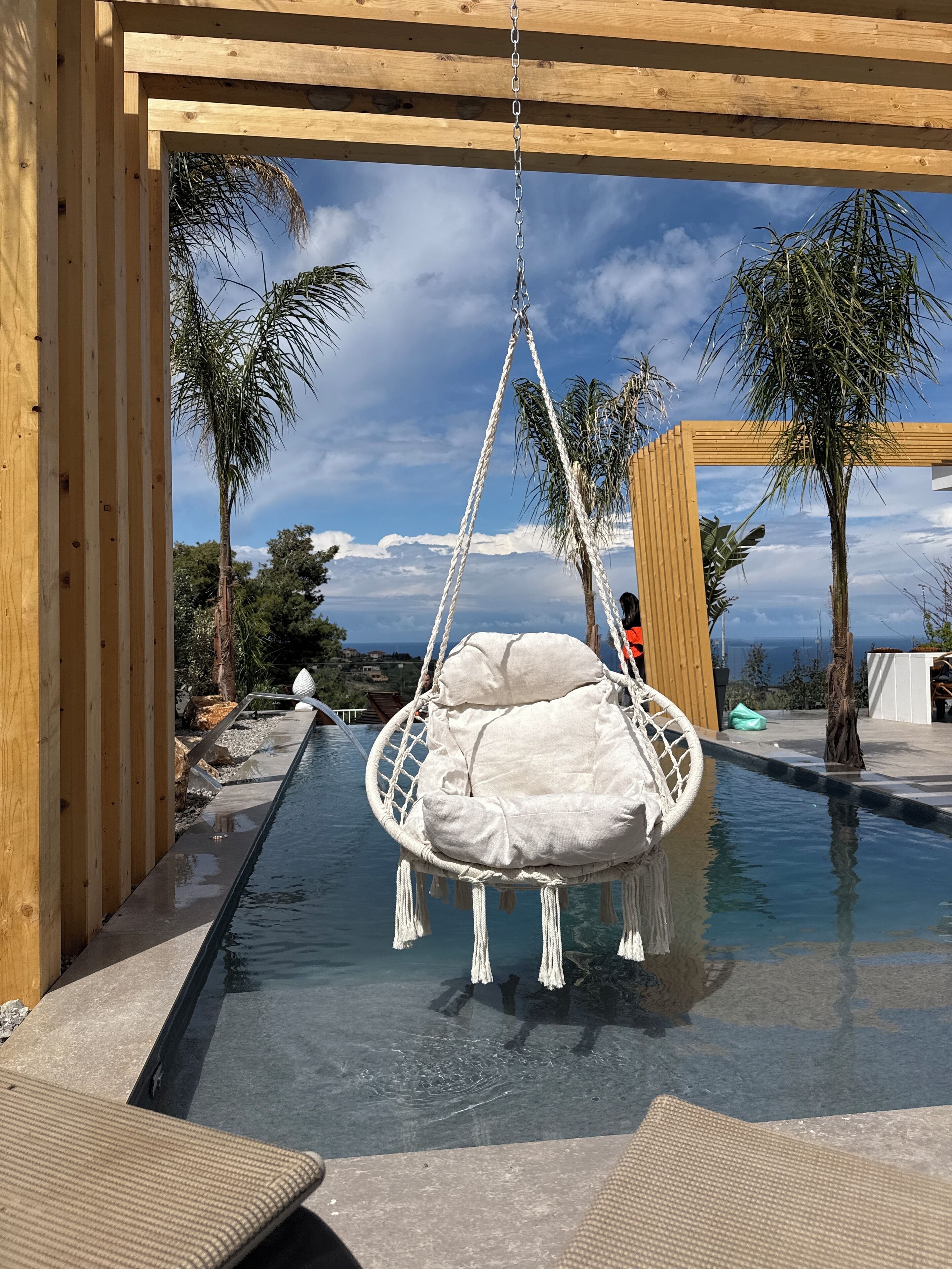 Une pergola en bois surmontée d’un fauteuil suspendu en forme de hamac blanc, au-dessus d’une piscine, le tout entouré de palmiers et sous un ciel dégagé.