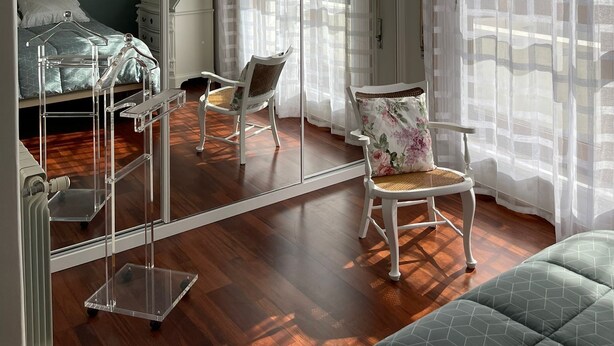 A bedroom with a large mirror, a hanging light fixture, a bed, a chair, and sheer curtains.