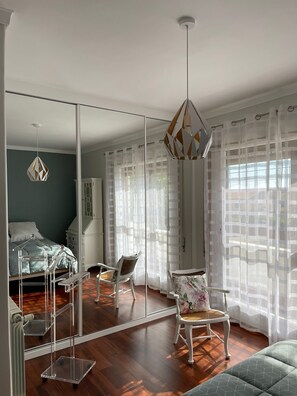 A bedroom with a large mirror, a hanging light fixture, a bed, a chair, and sheer curtains.