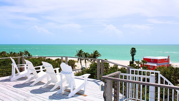 Une terrasse avec des fauteuils Adirondack blancs donnant sur une plage, avec un bâtiment rouge en arrière-plan.