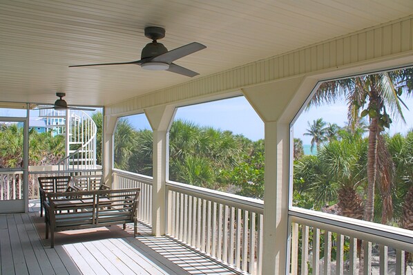 A covered outdoor area with a wooden deck, a bench, a ceiling fan, and large windows offering views of palm trees and a building in the background.