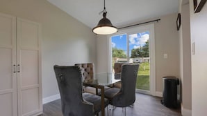 Een eetruimte met een glazen tafel en vier stoelen, een hangende lamp en uitzicht op een achtertuin door een schuifpui.