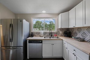 A compact kitchen with a stainless steel refrigerator, a dishwasher, a sink with a modern faucet, and a window with a view of greenery outside.