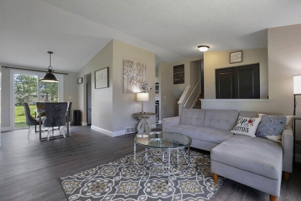A spacious living room with a grey sofa, a glass coffee table, and a patterned rug.