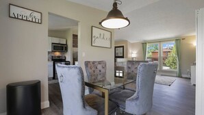 Een eetgedeelte met een glazen tafel en grijs gestoffeerde stoelen, een keuken op de achtergrond en een bord op de muur met de tekst 'gather'.