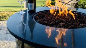 Una mesa con una botella de vino y una copa, un hogar, y una silla con cojín, con palmeras y un campo de golf al fondo.