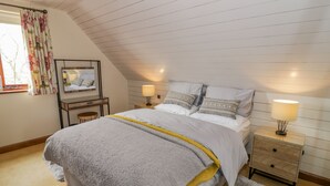 A bedroom with a sloped ceiling, a bed with white bedding, two bedside lamps, a wooden nightstand, a mirror, and a window with floral curtains. - The Stables (Stourbridge)