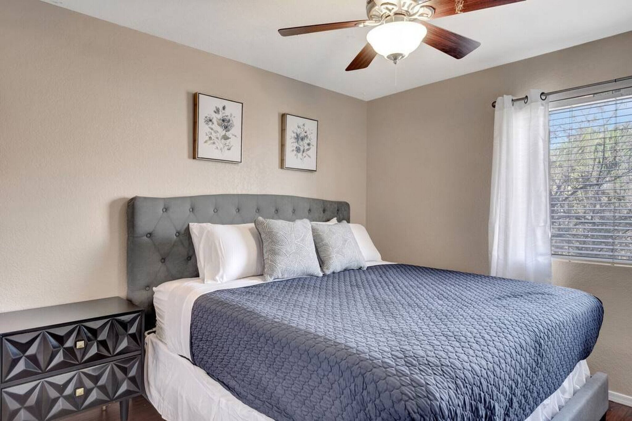 A bedroom with a tufted headboard, a bed with a blue quilt, a ceiling fan, and two framed floral artworks on the wall.