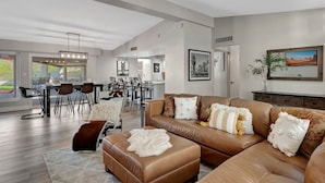 Un salon spacieux avec un canapé en cuir marron, un coin repas doté d’une table et de chaises modernes, et une grande fenêtre offrant une vue sur l’extérieur.
