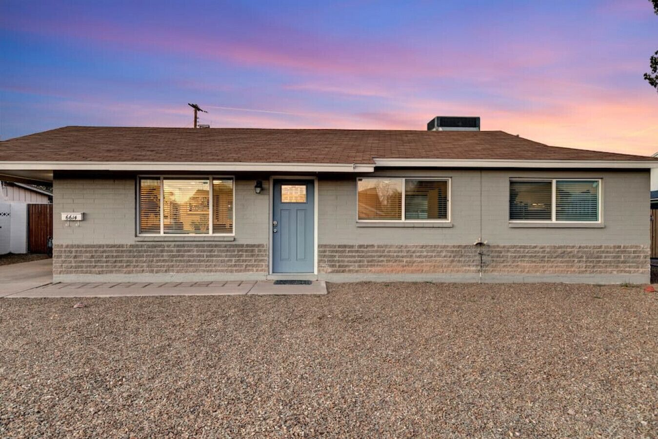 A single-story house with a blue door, two windows with blinds, and a gravel driveway.