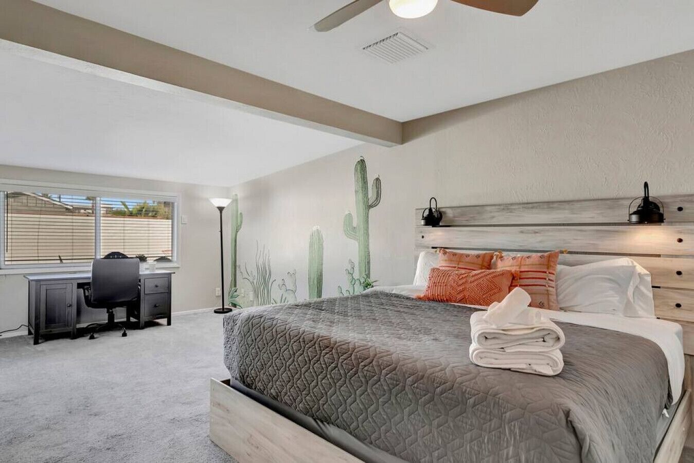 A bedroom with a bed, a desk, and a chair. There is a window with blinds, a cactus wall decoration, and a ceiling fan.