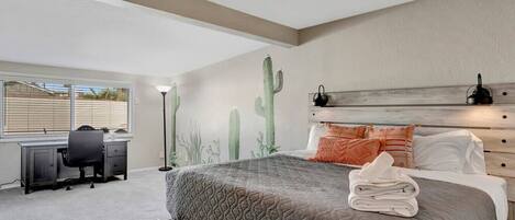 A bedroom with a bed, a desk, and a chair. There is a window with blinds, a cactus wall decoration, and a ceiling fan.