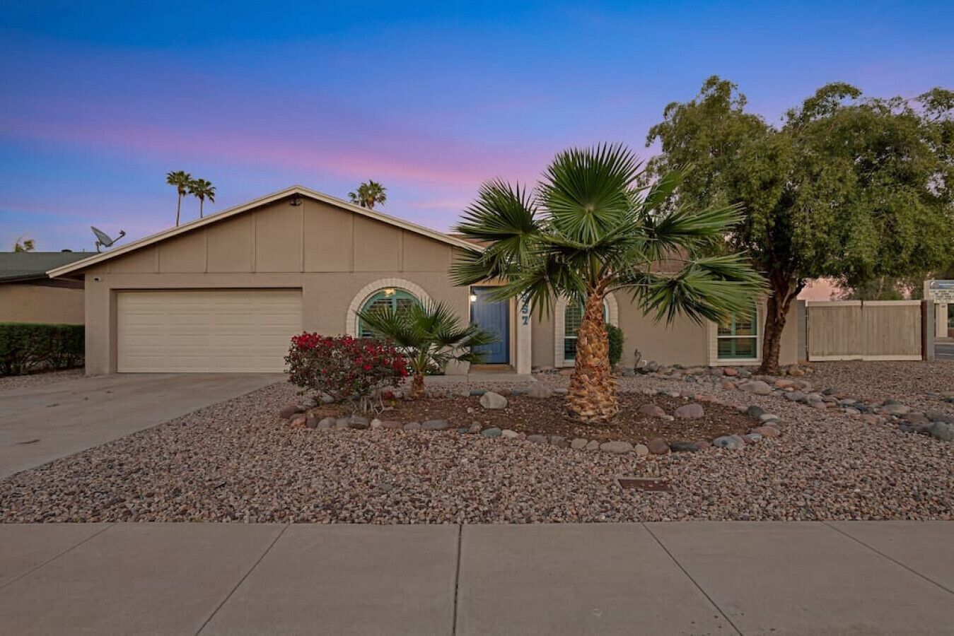 A residential house with a garage, a driveway, and a landscaped front yard with a palm tree and red flowers.