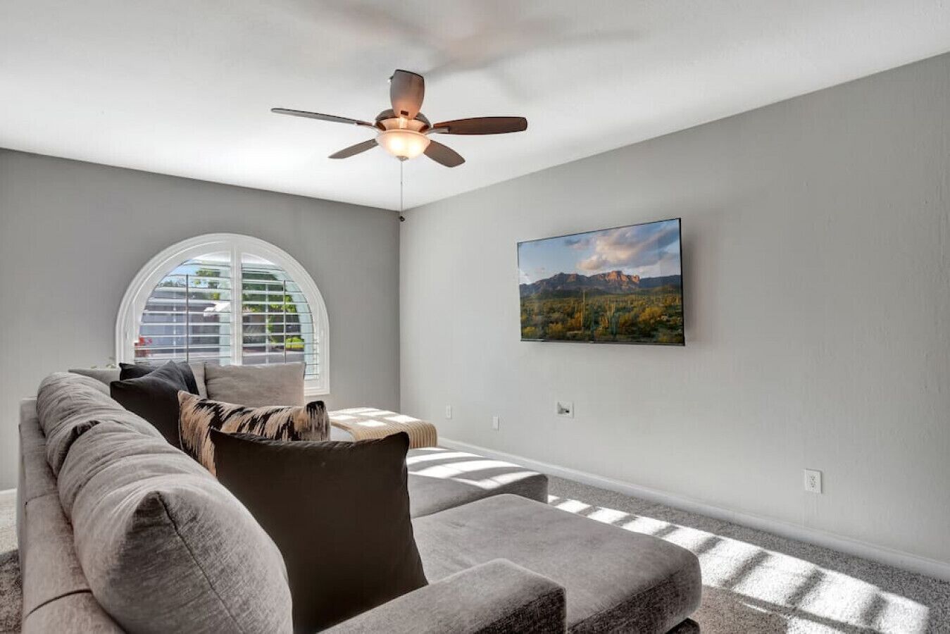 A living room with a grey sofa, a ceiling fan, a large window, and a framed picture on the wall.