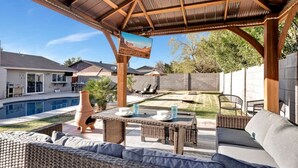 Une terrasse extérieure couverte, aménagée avec des meubles en osier, une piscine et une vue sur les maisons voisines.