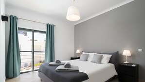 Quarto moderno com uma cama grande, duas mesas de cabeceira com luminárias, vista por uma porta de correr e uma varanda.