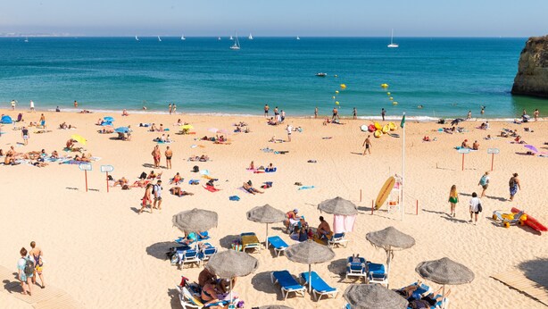 Uma praia com espreguiçadeiras e guarda-sóis, céu claro e veleiros ao longe.