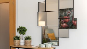 Espelho fixado na parede com moldura geométrica, uma mesinha com plantas em vasos e um quadro de flamingos.