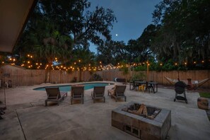 A backyard with a swimming pool, lounge chairs, a fire pit, and string lights.