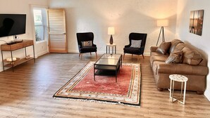 Ein Wohnzimmer mit Flachbildfernseher, zwei Sesseln, einer Couch, einem Couchtisch, einem Teppich und einem Deckenventilator.