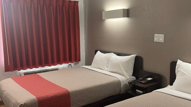 Een hotelkamer met twee bedden, elk met een rode en beige sprei, een nachtkastje met een telefoon, en een wandlamp.