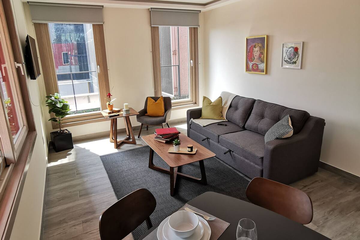 A modern living room with a grey sofa, a dining table set with plates and cutlery, a small coffee table, and a potted plant.