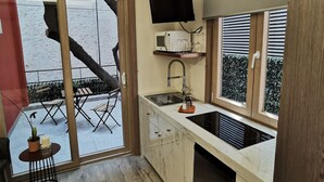 Eine moderne Küche mit integriertem Mikrowellenherd, Spüle und Herd. Ein großes Fenster mit Jalousien, ein flachbildschirmfernseher, der über der Arbeitsfläche angebracht ist, und eine kleine Außenlounge sind durch die Glastüren sichtbar.