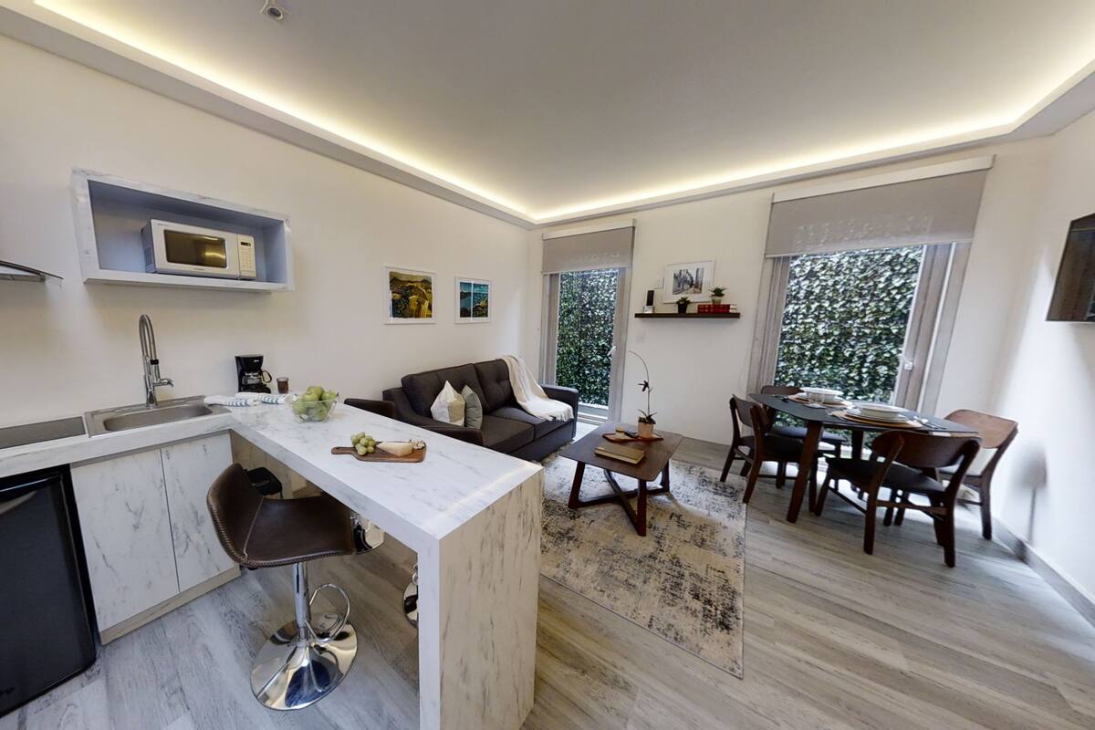 A modern kitchen with a built-in microwave, a coffee machine, and a sink. There is a dining area with a table and chairs, and a sofa.