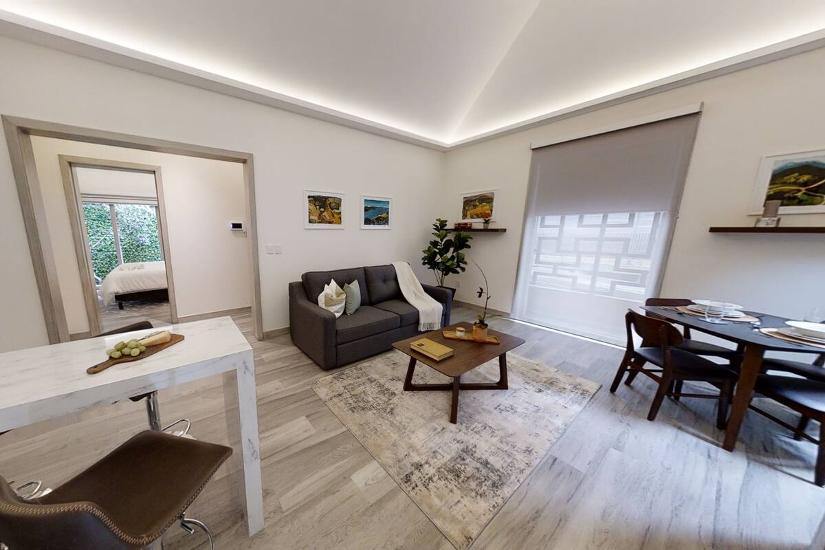 A modern living room with a sofa, coffee table, and dining area. There is a bed visible through an open door.