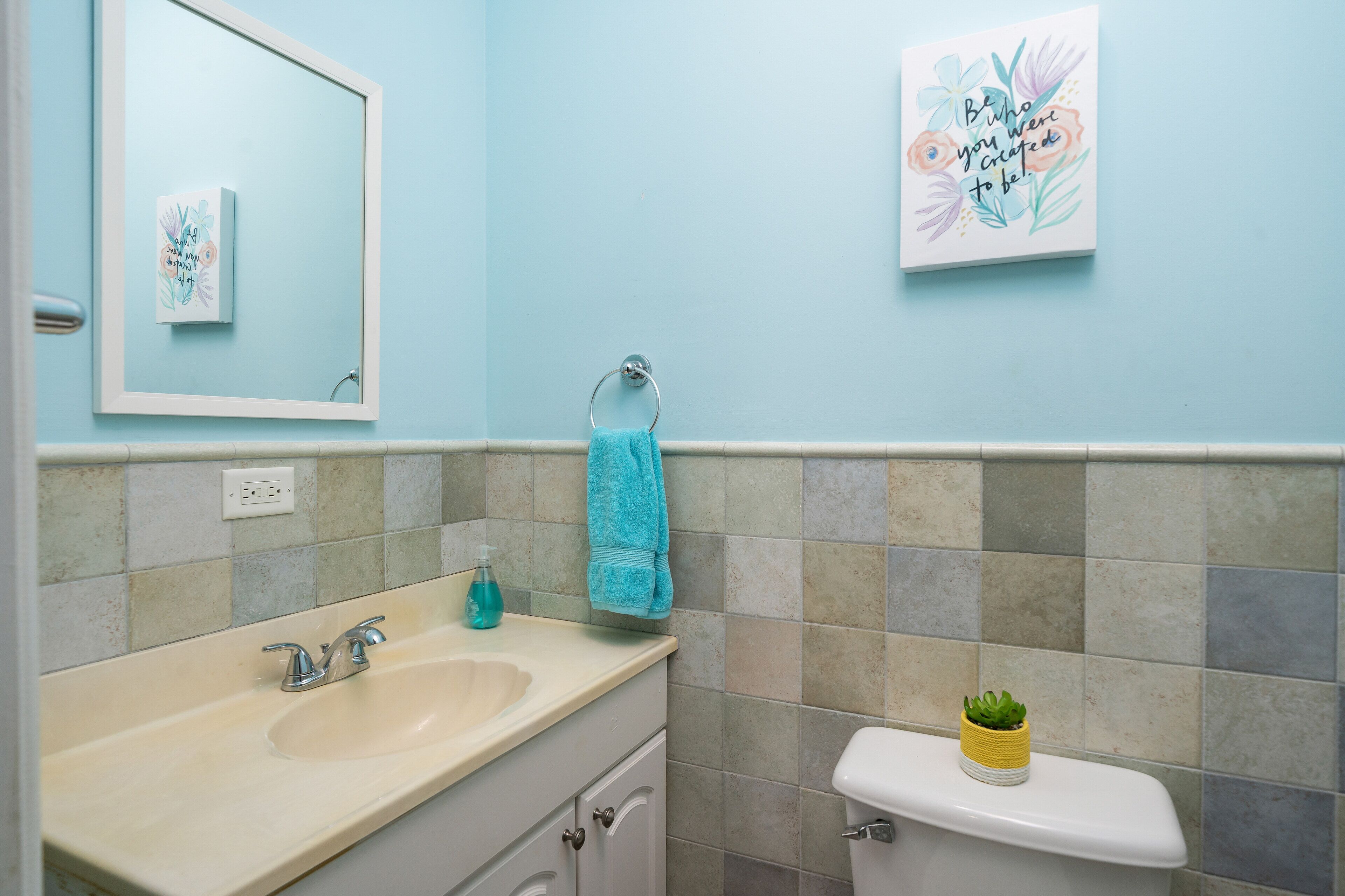 Ein Badezimmer mit Waschbecken, Spiegel und Toilette. Zwei gerahmte Kunstwerke sind an der Wand angebracht.