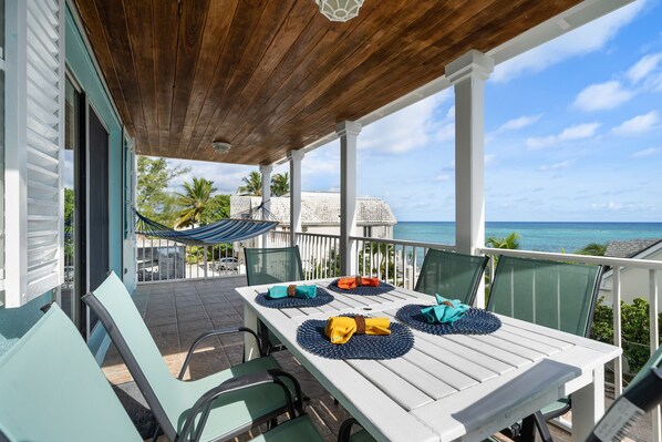 A covered outdoor dining area with a view of the ocean, white table, green chairs, and a hammock.