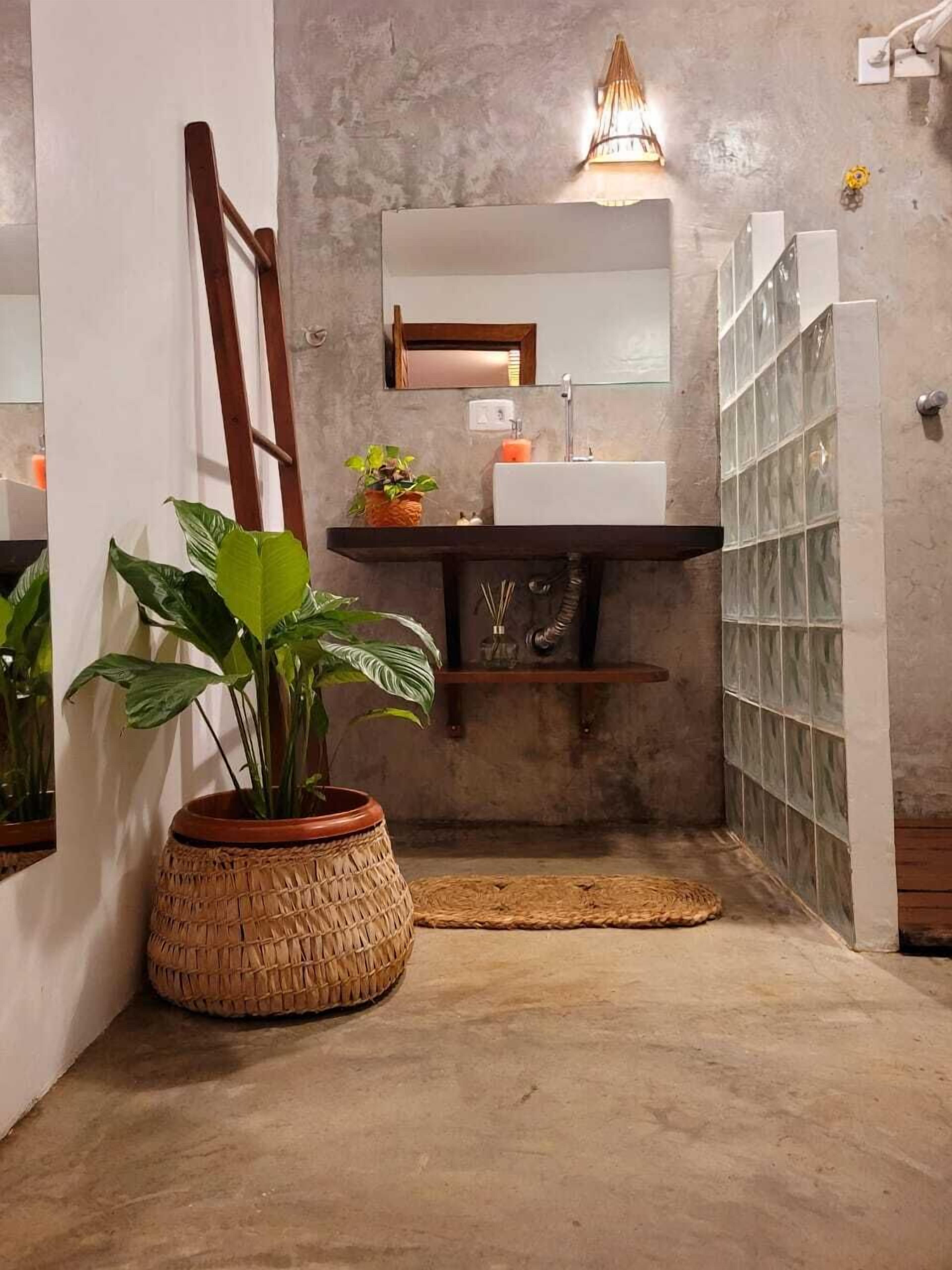 Une salle de bain avec un sol en béton, une échelle en bois appuyée contre le mur, un panier tressé contenant une plante, un lavabo blanc, un miroir et une cabine de douche en verre.