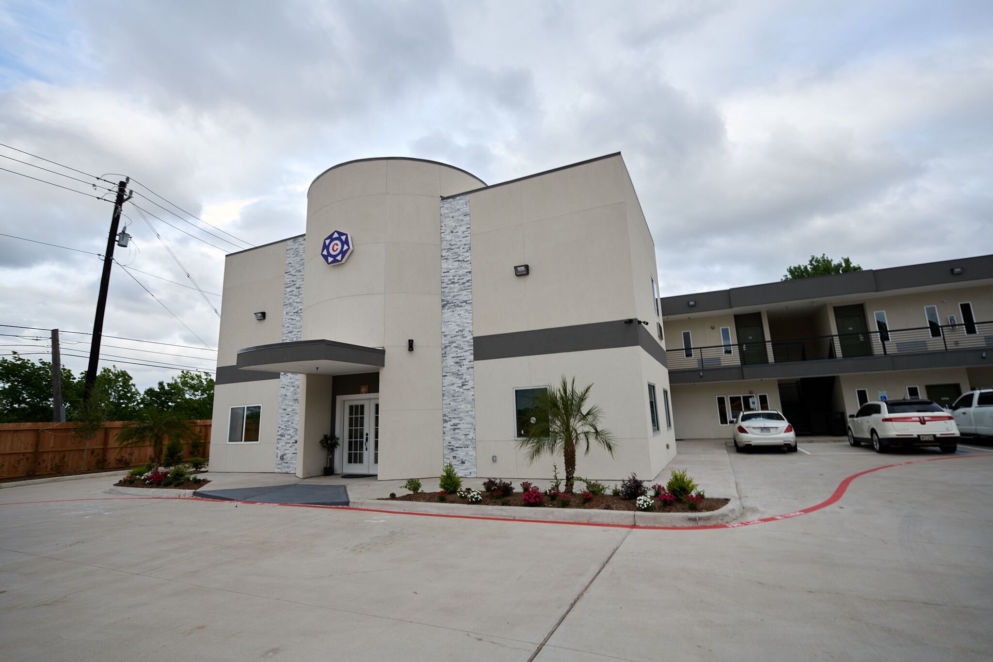 A modern building with a curved facade, a prominent logo, and a parking area with several cars.