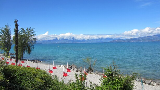 A beach with red umbrellas and white poles, a clear blue sky, and mountains in the background.