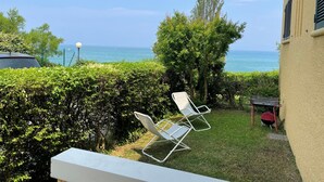A balcony with two white chairs facing a view of the sea, a car parked nearby, and a small table with a red chair.